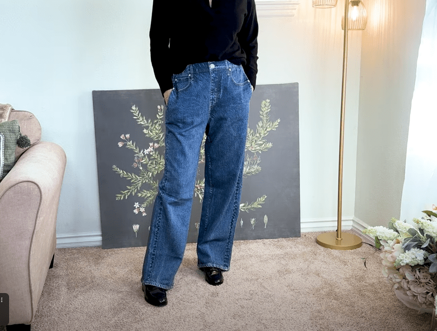 A person exudes effortless style and comfort wearing a black top and blue wide-leg jeans, standing with hands in pockets. The room boasts a pink couch, a tall lamp, a floral painting on the wall, and a bouquet on the floor.